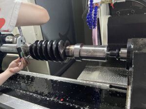 A close-up view on the SMK1507 CNC Thread Grinding Machine of a large black worm gear held between centers, being measured by an operator with a digital micrometer. The grinding wheel is in the background.