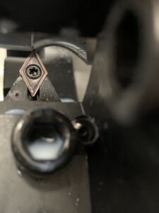 Macro photography of the whirling process, showing a diamond-shaped carbide insert engaging with the workpiece. The focus is on the cutting edge, illustrating the high-precision metal removal characteristic of CNC thread whirling.