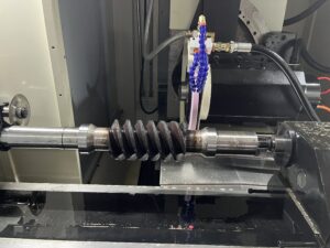 An operational shot from above of the pink grinding wheel in contact with a worm gear on the SMK1507 CNC Thread Grinding Machine. Coolant is actively spraying from blue multi-nozzles onto the grinding zone.